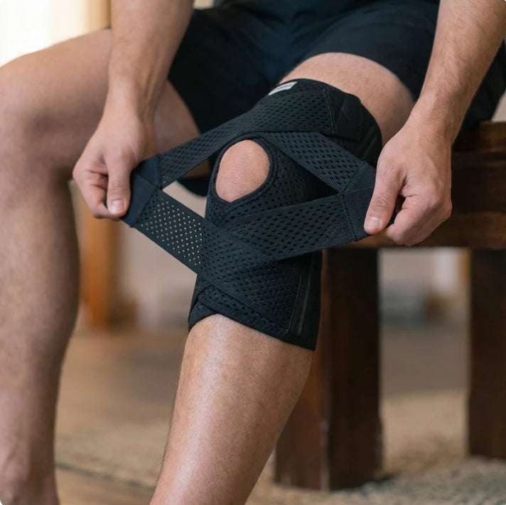 A person sitting on a wooden bench wearing a black knee immobilizer brace and adjusting the straps around their knee.